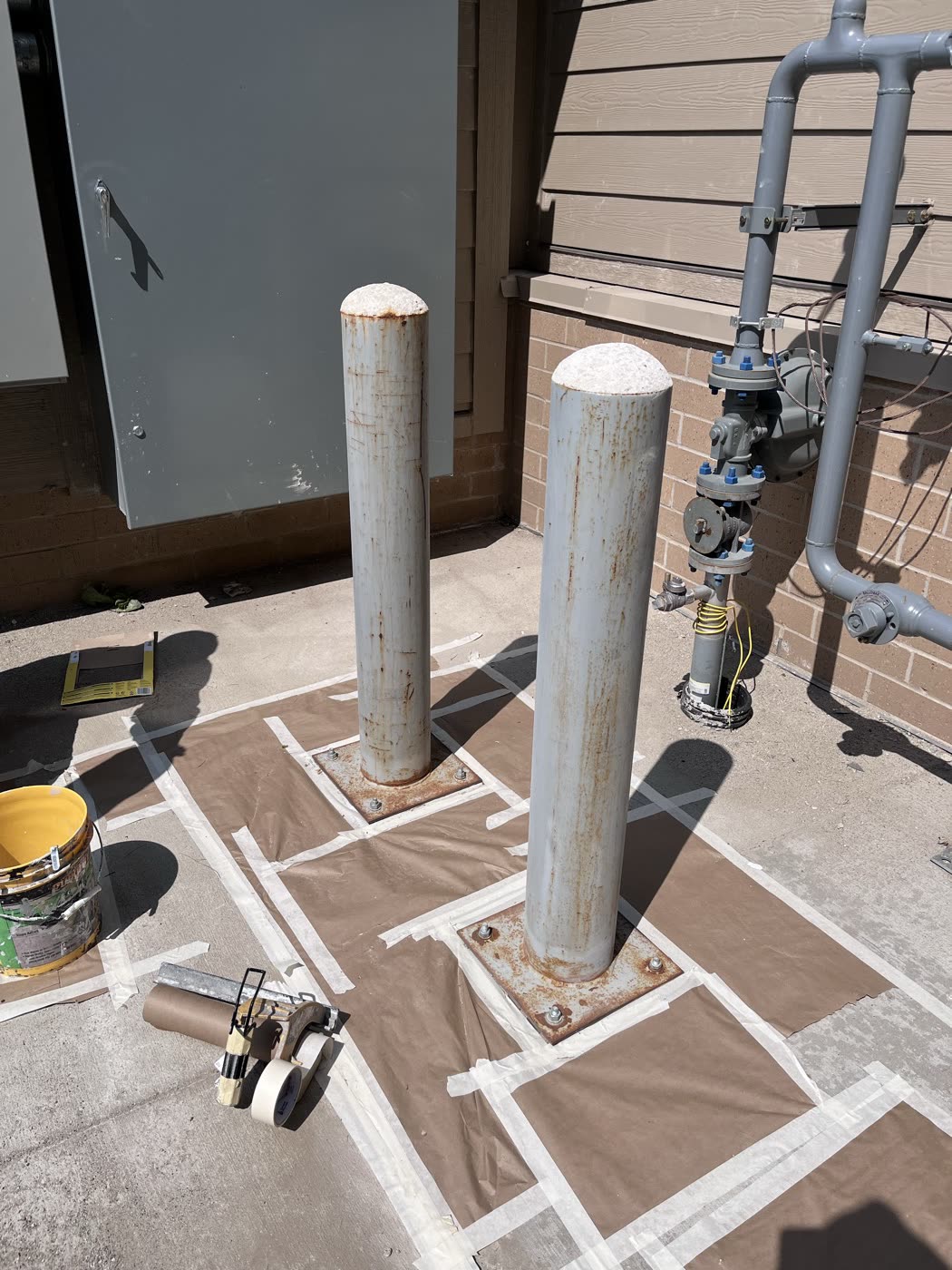 Two gray posts on a concrete pad with the ground masked off and a can of yellow paint nearby
