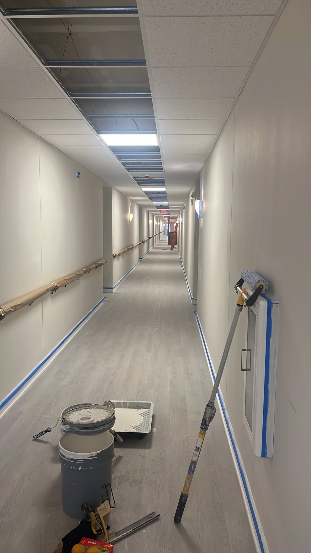Long hallway with taped baseboards, a paint roller and paint supplies on the floor